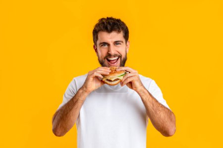 Photo for Junk food eater man biting delicious takeaway cheeseburger smiling to camera, standing on yellow backdrop in studio, enjoying fastfood meal, portrait shot. Diet vs overeating habit - Royalty Free Image