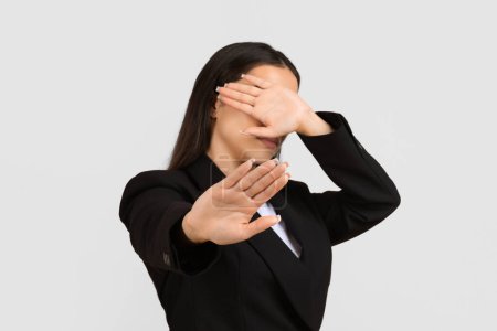 Professional woman in black blazer making stopping gesture with her palm while simultaneously covering her face, signaling rejection or avoidance