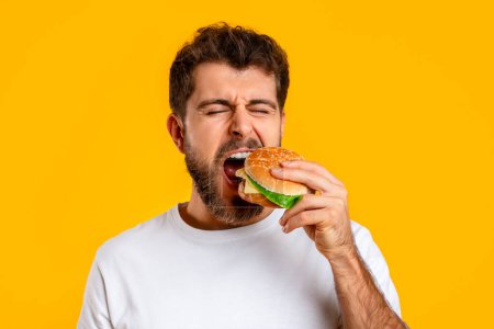 Foto de Divertido hombre caucásico toma bocado de tentadora hamburguesa en el fondo amarillo en el estudio, primer plano retrato de barbudo millennial consumiendo comida chatarra con apetito. Concepto de comida rápida - Imagen libre de derechos