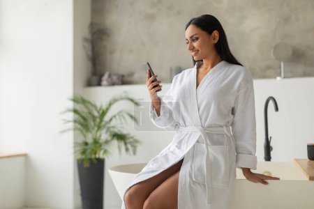 Téléchargez les photos : Attrayant jeune femme indienne aux cheveux longs en peignoir blanc assis sur la baignoire dans la salle de bain, lire un blog de beauté ou regarder des vidéos en ligne, à l'aide d'un smartphone, copier l'espace - en image libre de droit