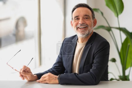 Photo for Charismatic happy handsome caucasian senior businessman with a beaming smile, holding eyeglasses and seated at a modern office table, with lush greenery in the background - Royalty Free Image