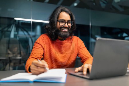 Téléchargez les photos : Homme hindou barbu joyeux dans occasionnel engage avec son ordinateur portable, prendre des notes dans un bloc-notes, son sourire reflétant la satisfaction et la productivité dans un espace de travail élégant. Entrepreneuriat, entreprise - en image libre de droit