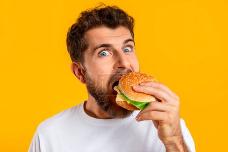 Photo for Portrait of funny european guy satisfying food cravings with cheeseburger, on yellow studio background, closeup. Millennial man taking big bite of tasty burger. Concept of cheat meal - Royalty Free Image