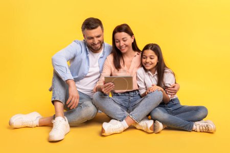 Photo for Family of three poses with digital tablet websurfing while sitting together on studio floor, over yellow background, representing modern e-learning or video call applications - Royalty Free Image