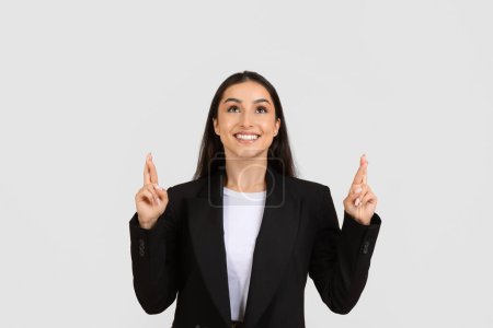 Photo for Optimistic young professional woman in black suit, crossing her fingers for good luck, looking up at free space with expectant smile, standing against plain background - Royalty Free Image