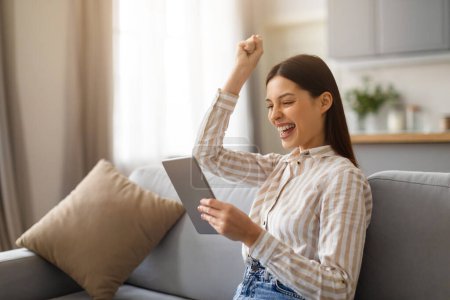 Photo for Elated young woman with beaming smile, triumphantly raising her arm while looking at tablet, resting sitting on cozy couch in bright living room, free space - Royalty Free Image