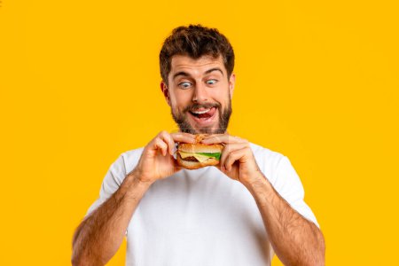 Téléchargez les photos : Homme barbu affamé drôle regarde burger avec tentation, se livre à cheeseburger incarnant l'envie de restauration rapide, portrait sur fond de studio jaune. Farine de cheat, nutrition - en image libre de droit