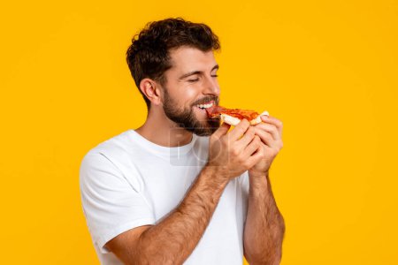 Téléchargez les photos : Portrait de jeune européen tient et savoure savoureux tranche de pizza, posant avec la malbouffe préférée en studio avec fond jaune, mettant l'accent sur le plaisir de profiter de la restauration rapide - en image libre de droit
