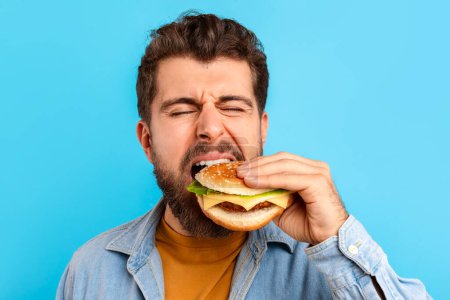 Photo for Funny bearded man enjoying cheat meal savoring burger against blue studio background, closeup portrait. Guy biting delicious fast food, tasting takeaway cheeseburger with appetite - Royalty Free Image