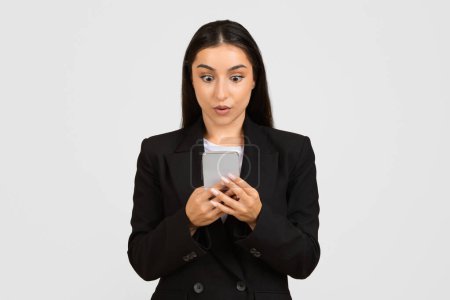 Photo for Perplexed young businesswoman in dark suit, eyes wide with surprise, intensely gazes at her smartphone screen, reacting to unexpected or astonishing information - Royalty Free Image