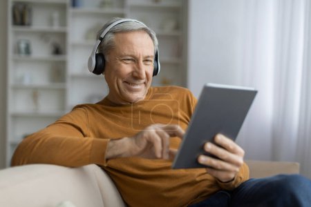 Hombre europeo de pelo gris positivo sentado en el sofá en casa, usando auriculares inalámbricos y almohadilla digital, viendo contenido de video, tener lección en línea, asistir a un seminario web