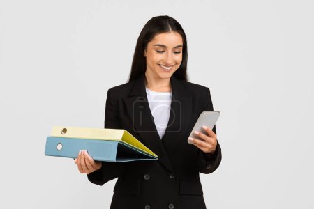 Engaged and efficient young businesswoman smiling while holding colorful folders and using smartphone, representing multitasking in professional setting