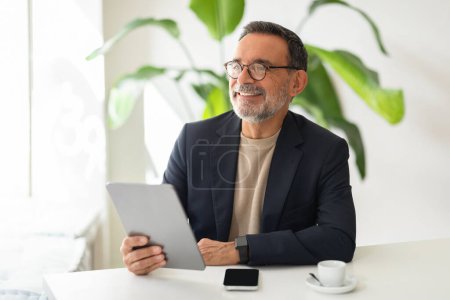 Photo for A contented caucasian senior businessman in a stylish navy blazer and beige turtleneck smiles while using a digital tablet in a modern, bright office with plants. Work, business app - Royalty Free Image