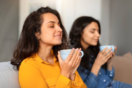 Téléchargez les photos : Deux jeunes femmes appréciant l'arôme de leur café tenant des tasses et fermant les yeux, assises dans un salon moderne paisible, se relaxant tout en buvant du thé chaud, vue latérale, foyer sélectif - en image libre de droit