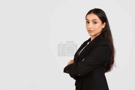 Téléchargez les photos : Jeune femme d'affaires professionnelle aux cheveux longs, vêtue d'un élégant costume noir, se tient avec confiance les bras croisés sur fond gris clair, espace libre - en image libre de droit