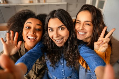 Téléchargez les photos : Trois jeunes femmes multiculturelles joyeux prenant selfie ludique, agitant et montrant des signes de paix, comme ils posent ensemble, faire du contenu de médias sociaux à la maison. Beauté et amitié diverses - en image libre de droit
