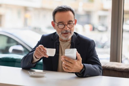 Téléchargez les photos : Homme caucasien senior en costume d'affaires profite d'une tasse d'espresso tout en naviguant sur son smartphone, un mélange de technologie moderne et de pause café traditionnelle. Travail, app entreprise - en image libre de droit