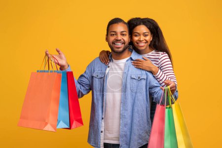 Photo for Happy Customers. Portrait Of Young Black Couple With Colorful Shopping Bags Embracing And Looking At Camera, Cheerful African American Spouses Posing Over Yellow Studio Background, Copy Space - Royalty Free Image
