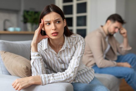 Photo for Concerned young woman in shirt holds her head, looking stressed, while upset man sits in the background with pensive expression, suggesting moment of personal or relational difficulty - Royalty Free Image