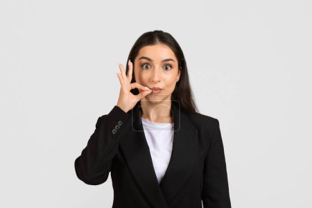 Photo for Engaging young businesswoman in sleek suit making quiet hand gesture with finger over her lips, showing playful secrecy on light grey backdrop - Royalty Free Image