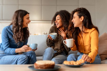 Téléchargez les photos : Trois heureuses femmes multiraciales parlent et rient, partageant leurs drôles d'histoires autour d'un café et d'un gâteau, assises sur un canapé dans un salon moderne à la maison. Véritable amitié, célébration chaleureuse - en image libre de droit