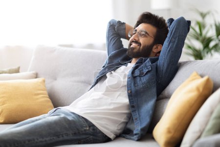 Photo for Home Rest. Smiling Dreamy Indian Guy Leaning Back On Comfortable Couch, Happy Eastern Man Relaxing On Sofa In Cozy Living Room Interior, Millennial Male Resting With Hands Behind Back - Royalty Free Image