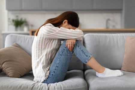 Photo for Distressed young woman in casual attire, sitting on sofa with her head resting on her knees, embodying posture of sadness, frustration or depression, side view - Royalty Free Image