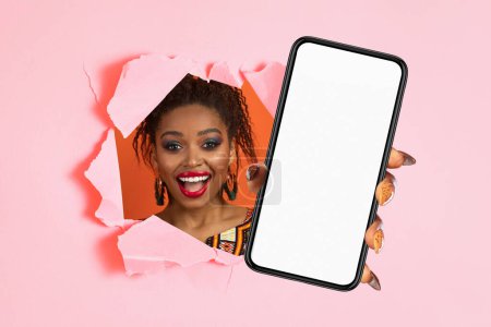 Glad afro-américaine jeune femme avec expression joyeuse et tenue traditionnelle regardant à travers un papier rose déchiré, tenant un smartphone avec un écran vide, pour le shopping de mode