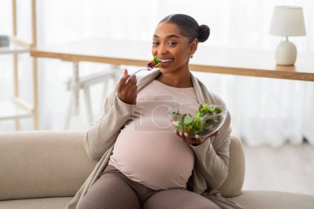 Photo for Cheerful young pregnant african american woman with big tummy sitting on couch at home, holding bowl with fresh organic meal, eating healthy salad. Diet, nutrition during pregnancy - Royalty Free Image