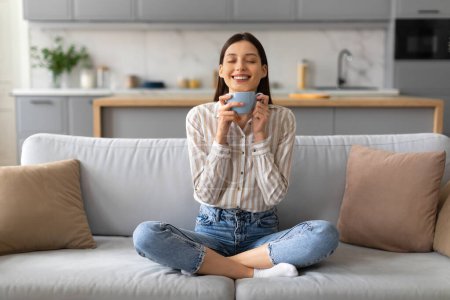 Photo for Cheerful young woman with radiant smile sits cross-legged on plush grey couch, savoring the warmth of blue mug of coffee in bright, modern living room - Royalty Free Image