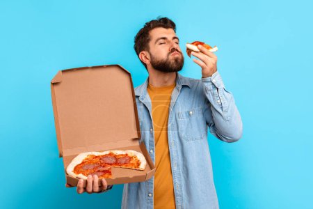 Photo for Young European man in casual attire eats juicy pizza slice while holds delivery box against blue backdrop, portrait of contented junk food eater savoring tasty meal. Comfort eating concept - Royalty Free Image