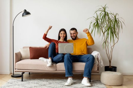 Photo for Happy Excited Young Couple Celebrating Success With Laptop At Home, Cheerful Millennial Spouses Looking At Computer Screen And Shaking Fists With Joy While Sitting On Couch In Living Room - Royalty Free Image