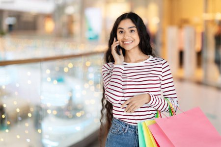 Photo for Emotional pretty long-haired young hindu woman wearing casual clothing walking by modern shopping mall, carrying colorful paper bags purchases, talking on phone, looking at copy space - Royalty Free Image