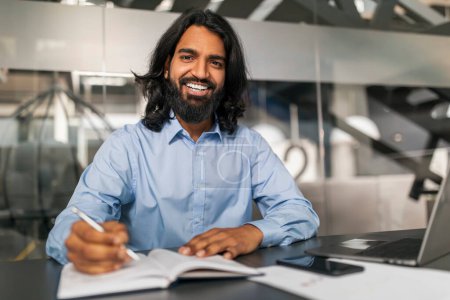 Photo for Positive bearded young indian man in formal employee working at modern office, sitting at desk with computer, taking notes in notebook, getting ready for presentation, online meeting, copy space - Royalty Free Image