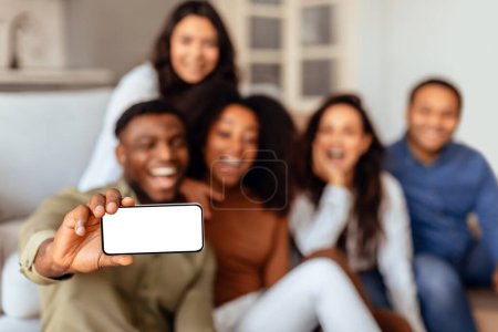 Foto de Grupo de diversos estudiantes amigos tomando selfie en el teléfono celular con pantalla en blanco, divertirse en fin de semana en la sala de estar en casa, haciendo contenido para las redes sociales durante la reunión. Burla, enfoque selectivo - Imagen libre de derechos