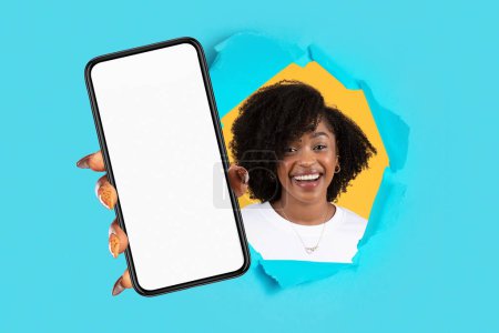 Foto de Sonriente dama afroamericana milenaria mostrando un teléfono inteligente con pantalla vacía a través de un agujero en papel, fondo de estudio azul. Sitio web, aplicación, recomendación, moda y venta - Imagen libre de derechos