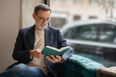 Photo for Thoughtful smart caucasian senior man with glasses engrossed in reading a book, comfortably seated near a window in a cozy corner with soft natural light, enjoy spare time - Royalty Free Image