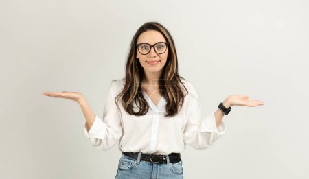 Photo for A young european woman with brown hair and glasses raises her hands with palms up in a shrugging pose, wearing a white blouse and jeans, on a light background, studio. Work, business - Royalty Free Image