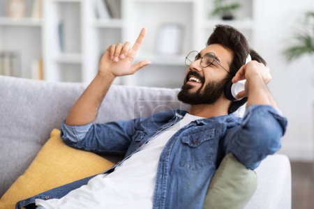 Téléchargez les photos : Positif jeune homme indien s'amuser tout en écoutant de la musique à la maison, gai gars de l'Est portant des écouteurs sans fil se détendre sur le canapé dans le salon, profiter de chansons préférées, gros plan - en image libre de droit