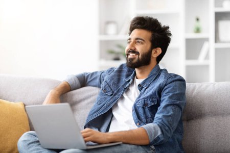 Photo for Portrait Of Young Smiling Indian Man With Laptop Relaxing On Couch At Home, Dreamy Millennial Eastern Guy Sitting With Computer On Sofa In Cozy Living Room And Looking Away, Copy Space - Royalty Free Image