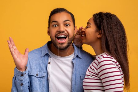 Photo for Big Secret. Young Black Woman Sharing News With Her Excited Boyfriend, Surprised African American Man Raising Hands In Amazement While Standing Together Over Yellow Studio Background, Free Space - Royalty Free Image