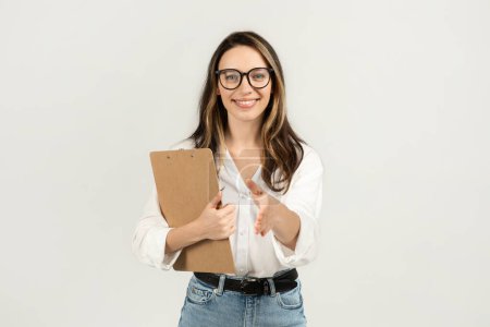 Téléchargez les photos : Accueillante jeune femme d'affaires européenne avec des lunettes tenant un presse-papiers et tendant la main pour une poignée de main, vêtue d'un chemisier blanc et d'un jean sur un fond simple, studio - en image libre de droit