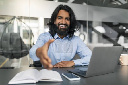 Photo for Cheerful bearded young indian business consultant outstretching hand towards camera and smiling, sitting at desk with laptop computer, smartphone and notebook at modern office, greeting client - Royalty Free Image