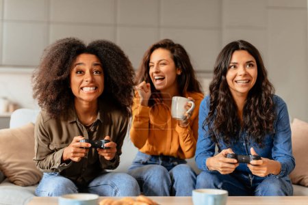 Téléchargez les photos : Des amies multiculturelles jouant à des jeux vidéo dans leur salon confortable, profitant d'une séance de jeu remplie de plaisir, riant engagées dans une compétition ludique, assises sur un canapé à la maison à l'intérieur - en image libre de droit