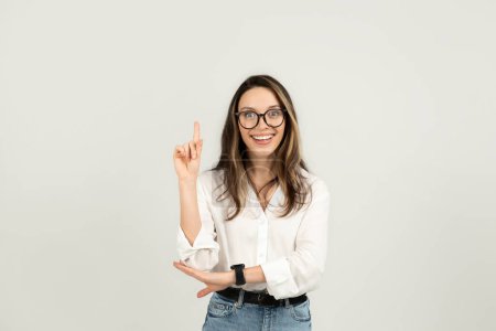 Foto de Mujer joven europea entusiasta con gafas apuntando alegremente hacia arriba, mostrando algo emocionante o teniendo una idea brillante, mientras está de pie con confianza, estudio. Negocios, trabajo - Imagen libre de derechos