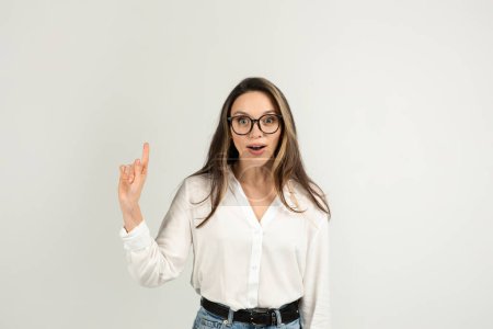 Foto de Una joven europea sorprendida con el pelo castaño largo y gafas grandes, vistiendo una blusa blanca y jeans, apunta hacia arriba con su mano derecha sobre un fondo liso, estudio - Imagen libre de derechos