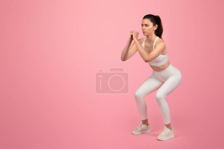 Photo for Focused caucasian woman in white fitness clothing performing a squat exercise, demonstrating strength, and concentration, promoting an active and healthy lifestyle on a pink background - Royalty Free Image