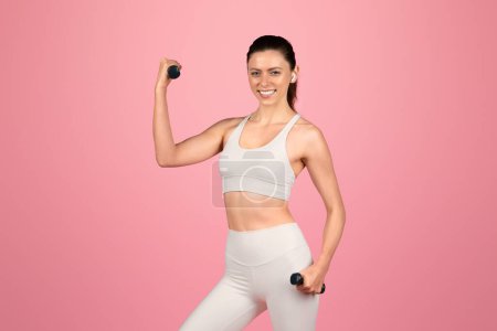 Téléchargez les photos : Enthousiaste femme en forme européenne en tenue d'entraînement blanche s'exerçant joyeusement avec des haltères, affichant énergie et positivité dans sa routine de fitness sur un fond rose - en image libre de droit