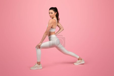 Photo for Focused and toned european young woman in a white sports bra and leggings performing a stretching exercise, showcasing strength and flexibility on a pink background, studio - Royalty Free Image