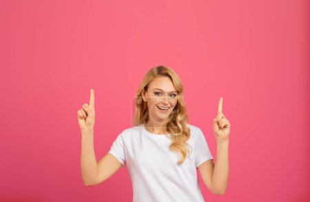 Glückliche junge blonde Frau im weißen T-Shirt, die mit beiden Händen nach oben zeigt, auf rosa Studiohintergrund. Millennial Lady zeigt ihre Empfehlung über dem Kopf. Freiraum